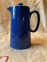 Front view of tall, deep blue ceramic coffee pot with handle and lid knob, set against a tan fabric background.