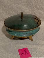 Right-side angled view of a vintage brass potpourri container with a green patina and tripod legs on light fabric.