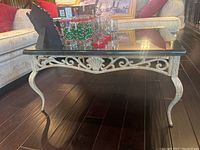 Front angle view of square coffee table showing glass top and ornate metal base with curved legs and shell motif.