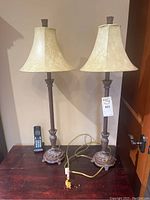 Pair of tall decorative vintage look bedside table lamps with beige bell-shaped shades and bronze bases, shown on a dark wood surface with cords visible and a cordless phone for scale.