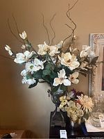Large artificial floral arrangement with white fabric flowers, green leaves, curly brown branches in metal urn vase shown against a beige wall