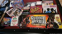 Boxed Guitar Hero 3 Legends of Rock for PlayStation 2 with wireless guitar and multiple boxed board games and puzzles on a table.