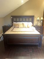 Front view of king size dark brown wooden bed with linens and pillows, set against slanted beige wall.