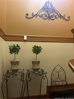 Photo showing two metal stands with two faux plants in decorative pots and metal hanging shelf with scroll accents on right side