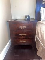 Two matching deep brown wooden nightstands each with three drawers and heavy metal handles. One nightstand shown alone, the other shown with lamps and electronics on top.