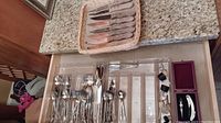 Silverware organized in a drawer including large and small spoons, forks, and steak knives in a basket on the counter.