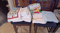Various folded table cloths and linens stacked on two chairs, showing white, colorful printed, and pastel blue fabrics.