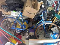 Full side view of a blue Raleigh 3 speed bicycle amidst clutter showing frame, tires, pedals, and rear rack.