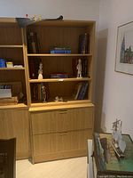 Full view of the wood bookcase standing against the wall, showing the three open shelves filled with books and figurines, and the two drawers at the bottom.