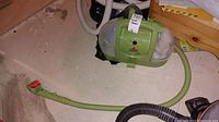 Angled photo showing the front and side of the green Bissell Little Green spot carpet cleaner with attached hose and cleaning nozzle laying on concrete floor.