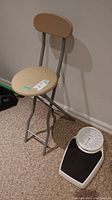 Photo of a metal folding chair with a faux wood round seat and backrest, standing on carpet next to a Seca floor scale.
