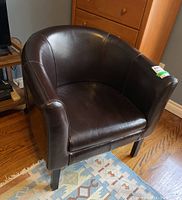 Front and angle view of the brown leather arm chair with curved back and armrests, situated on a hardwood floor with patterned rug.