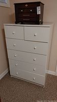 Front view of white laminate dresser with six drawers and a small dark wooden chest placed on top.