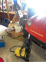 Full view of electric power washer showing the yellow body, black handle, hose, and spray wand leaning against a red car and storage items.
