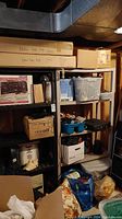 Two plastic utility shelving units located in a basement filled with various storage boxes and items. Black shelving unit is prominently visible with contents on shelves.