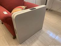 Portable folding table shown folded closed leaning against a chair, showing plastic beige textured surface and integrated handles.