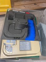 Dark gray plastic toy base plate with a mapped parking lot, ramp, and blue water feature, stacked with beige base plates having printed stone stair and floor designs inside a clear bin.