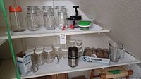 Various glass jars with white and metal lids on shelves including tall jar with orange lid, small green grater, hand-operated spice grinder, glass pitcher, and some small containers.