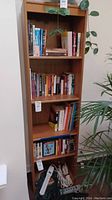 Full view of solid wood bookshelf filled with books, showing adjustable shelves, standing next to plants and a wall