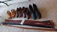 Three pairs of women's footwear in tan, brown, and black colors along with six assorted leather belts lying on a carpeted floor.