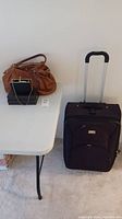 Full view of the brown purse, small black clutch, and black roller suitcase on the floor next to a white folding table.