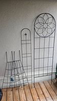 Photo showing four black metal garden trellises including the two identical smaller trellises in front and two larger trellises behind on wooden floor against wall.