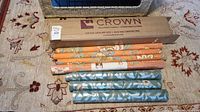Seven wallpaper rolls laid on carpet including three blue/green/silver William Morris style and four orange floral Jaima Brown Home rolls with one sealed in box
