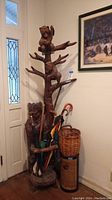 Front view of carved wood bear coat hanger with attached tree branch hooks, umbrella stand filled with umbrellas and walking sticks, alongside wicker basket and tin container.