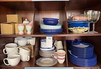 Wide view of shelves with stacked plates, cups, bowls, egg cups and plastic picnicware including plastic wine glasses