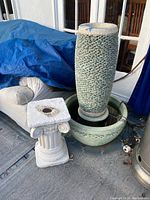 Wide shot showing tall pebble-textured fountain next to white decorative cement pillar, both outdoors on patio surface with some surrounding objects and blue tarp in background.