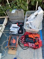 Full view of the lot showing gray Skil drill case open with cordless drill inside, a Bosch hard plastic case, a Black & Decker boxed hand sander, and a plastic bin holding a white Eufy vacuum with attachments.