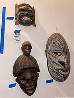 Three carved tribal masks mounted on a white wall; labels indicate sizes and notes about wood and ceramic composition