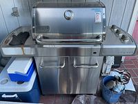Front view of large stainless steel Weber Genesis BBQ grill showing cabinet doors, side shelf with control knobs, and lid with handle.