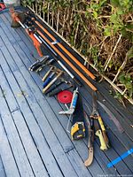 Photo showing an orange and black string trimmer and long-handled orange Fiskars pole tools (pruner and saw) placed on blue wooden deck beside green shrubbery.