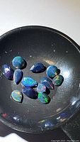 Photo of 13 calibrated pear cut Ethiopian black opals showing various vibrant play-of-color on a black surface under direct light.