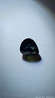 Pear-shaped sapphire gemstone with deep dark blue-green color placed against a white surface under light.
