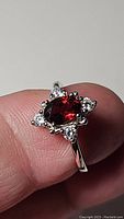 Close-up photo of the garnet ring showing the deep red oval garnet and clear accent stones on polished silver adjustable band, worn on finger for scale.