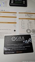 Photo of GRA Moissanite warranty card and certification report showing the 1.0ct weight, D color grade, and VVS1 clarity grade.