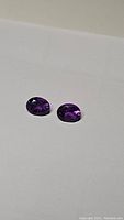 Pair of deep purple amethyst oval cut gemstones displayed on white background, showing clarity and color.