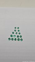 Top view of 15 natural calibrated emerald gemstones arranged in a pyramid shape showing different sizes and green shades.