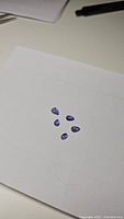 Five pear cut purple blue Tanzanite cabochons laid out on white paper, showing color and shape with visible inclusions.