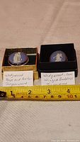 Photo showing both brooches side by side in open boxes with handwritten labels indicating design and jasperware origin.