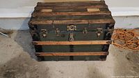 Closed antique trunk with green body, black metal reinforcements, and wooden slats.