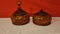Two carved wooden bowls with lids, showing floral carved patterns and different lid finials, against a red background.