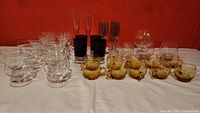 The full lot on a white surface showing various vintage glassware items including clear stemmed glasses, amber small cups, tall dark amber glasses, and champagne flutes.