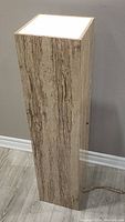 Tall rectangular light up art base in faux stone or wood grain finish, top illuminated panel glowing while lit, showing full side view with power cord.