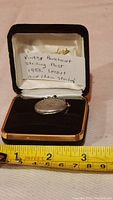 Photo showing the sterling silver locket and chain inside an open jewelry box with a note indicating 'Vintage Buckhardt Sterling Post 1953 locket and chain Sterling'. A measuring tape indicates the approximate size of the locket.