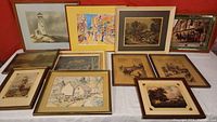 Overview photo showing 11 framed prints and artworks arranged on a surface against a red background wall, variety of styles and subjects.