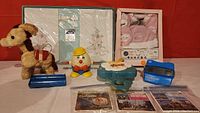 Wide shot showing the full lot including the stuffed pull toy dog, Humpty Dumpty toy, blue stapler, Fisher-Price piano toy, packaged pink baby clothes, packaged floral sheet set, and View-Master reel sets.