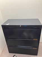 Front view of a 3 drawer black metal filing cabinet with lock and key inserted in the top drawer lock.